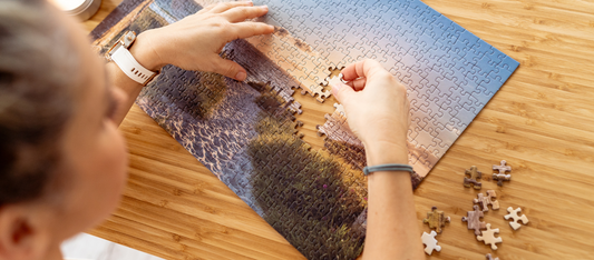 Person legt ein Puzzleteil in ein Landschaftspuzzle mit Meer, Felsen und Himmel auf einem Holztisch