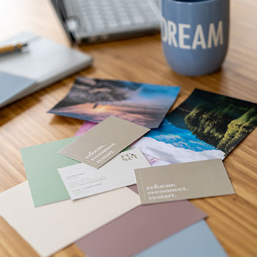 Verschiedene Postkarten und Farbmuster auf einem Holztisch, im Hintergrund eine Tasse mit der Aufschrift 'DREAM' und ein Laptop
