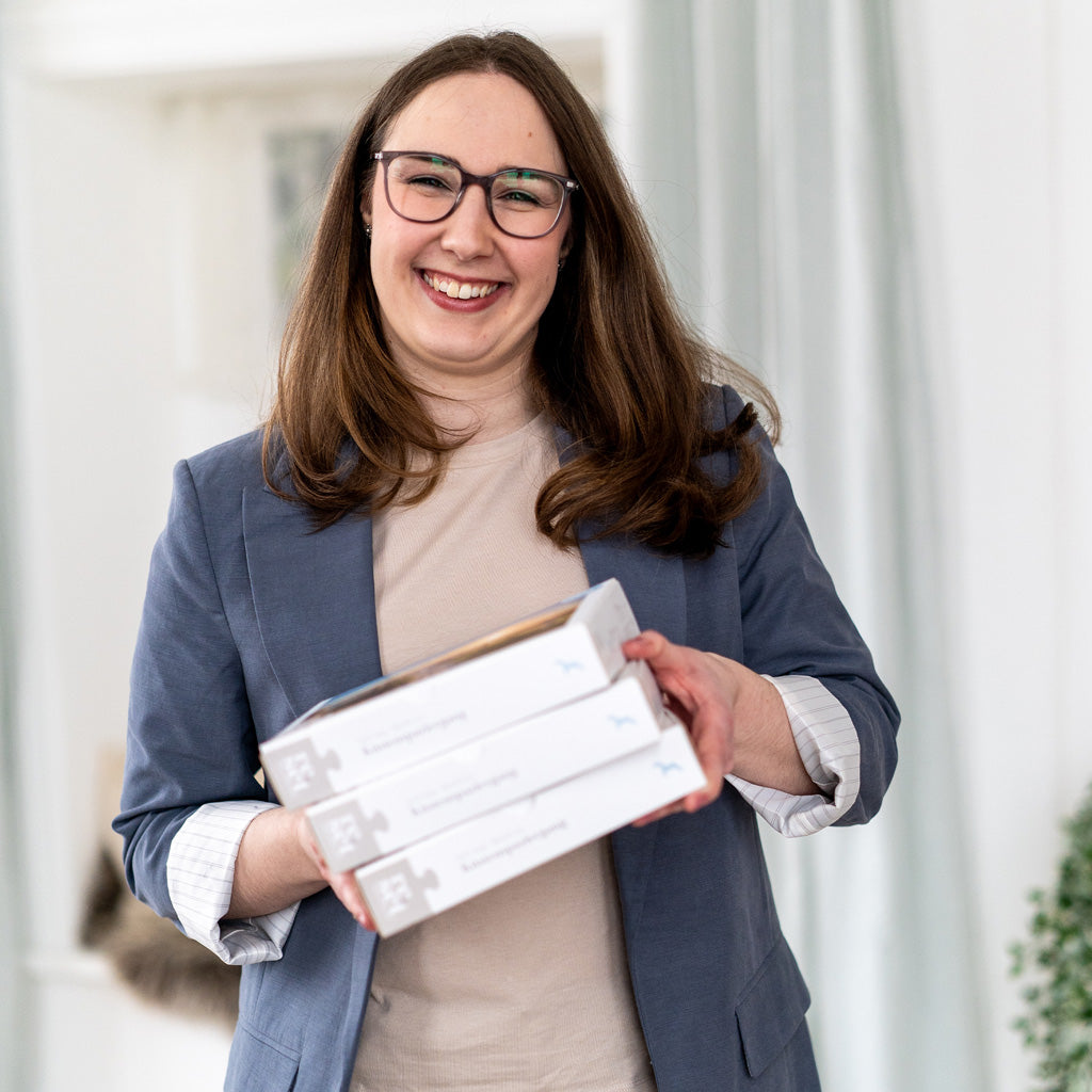 Lächelnde Gründerin des Unternehmens mit Brille und blauem Blazer hält drei hell-beige Produktverpackungen in den Händen