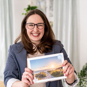 Lächelnde Frau mit Brille und dunklem Blazer hält eine Box mit einem Bild eines Sonnenuntergangs am Strand, Gründerin des Unternehmens