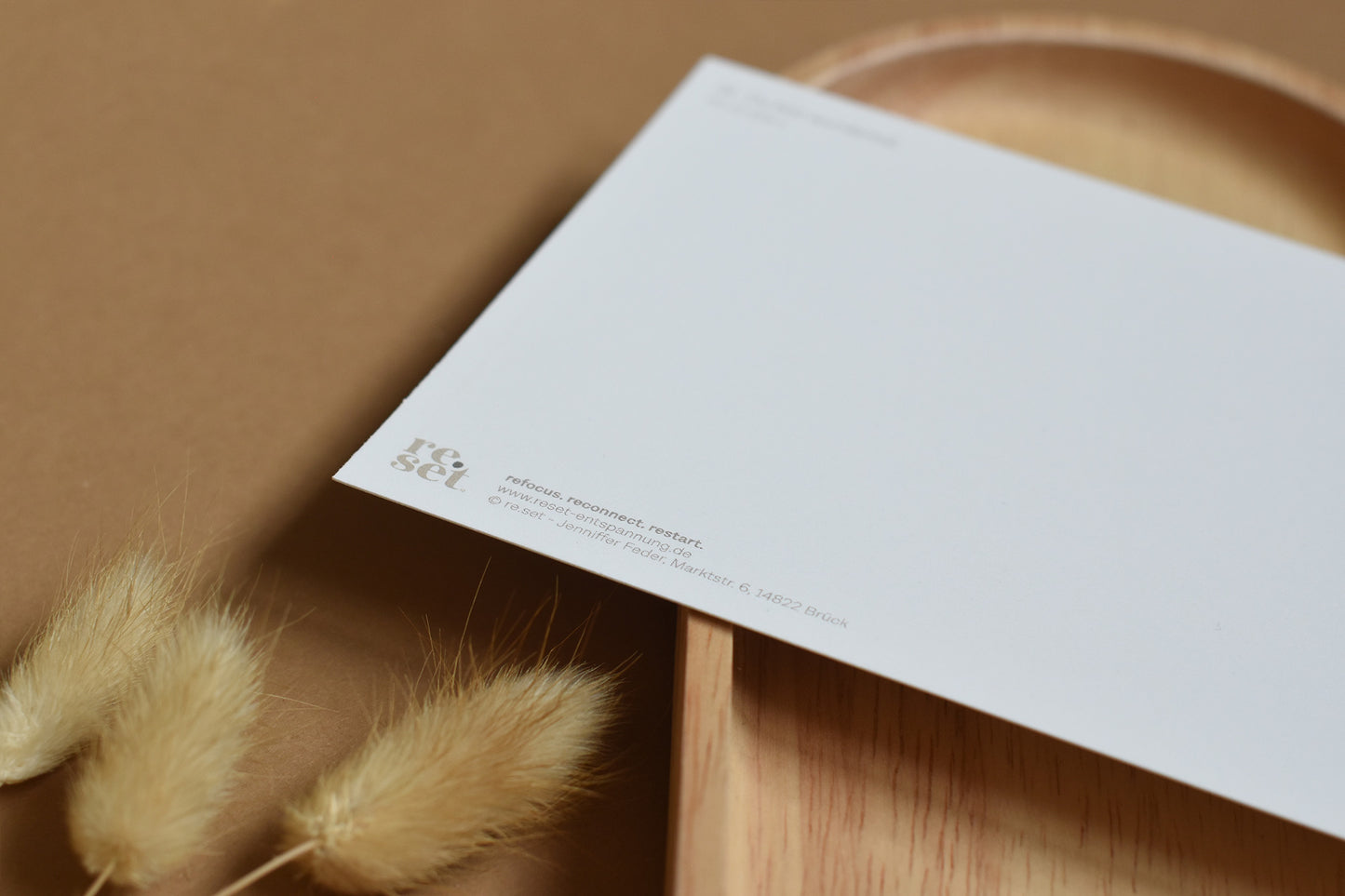 Rückseite einer weißen Postkarte mit kleinem Logo 'reset' auf braunem Untergrund mit getrockneten Gräsern und Holztablett