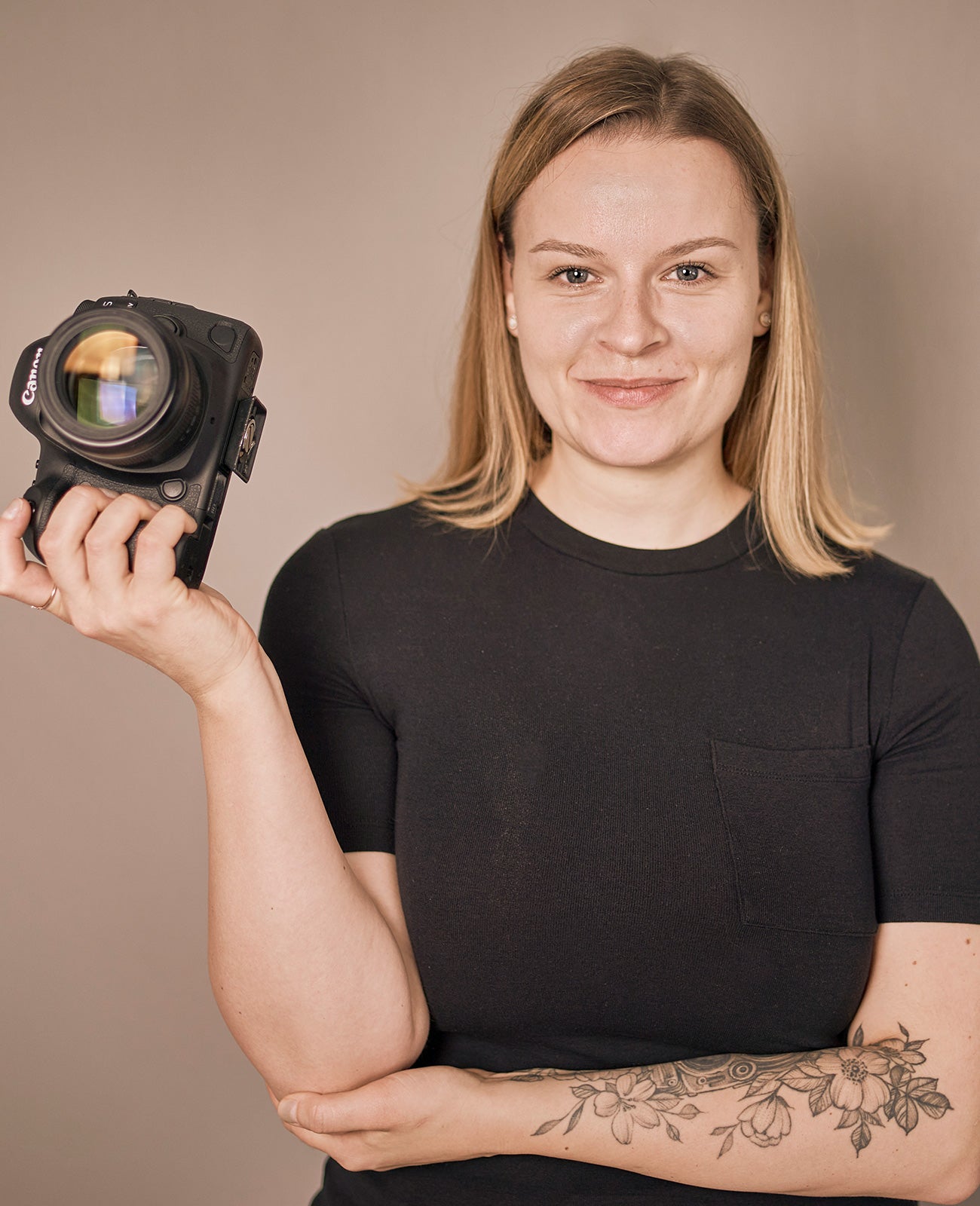Frau mit schulterlangen blonden Haaren und schwarzem T-Shirt hält eine Canon-Kamera in der rechten Hand, sichtbares Blumen-Tattoo am linken Unterarm