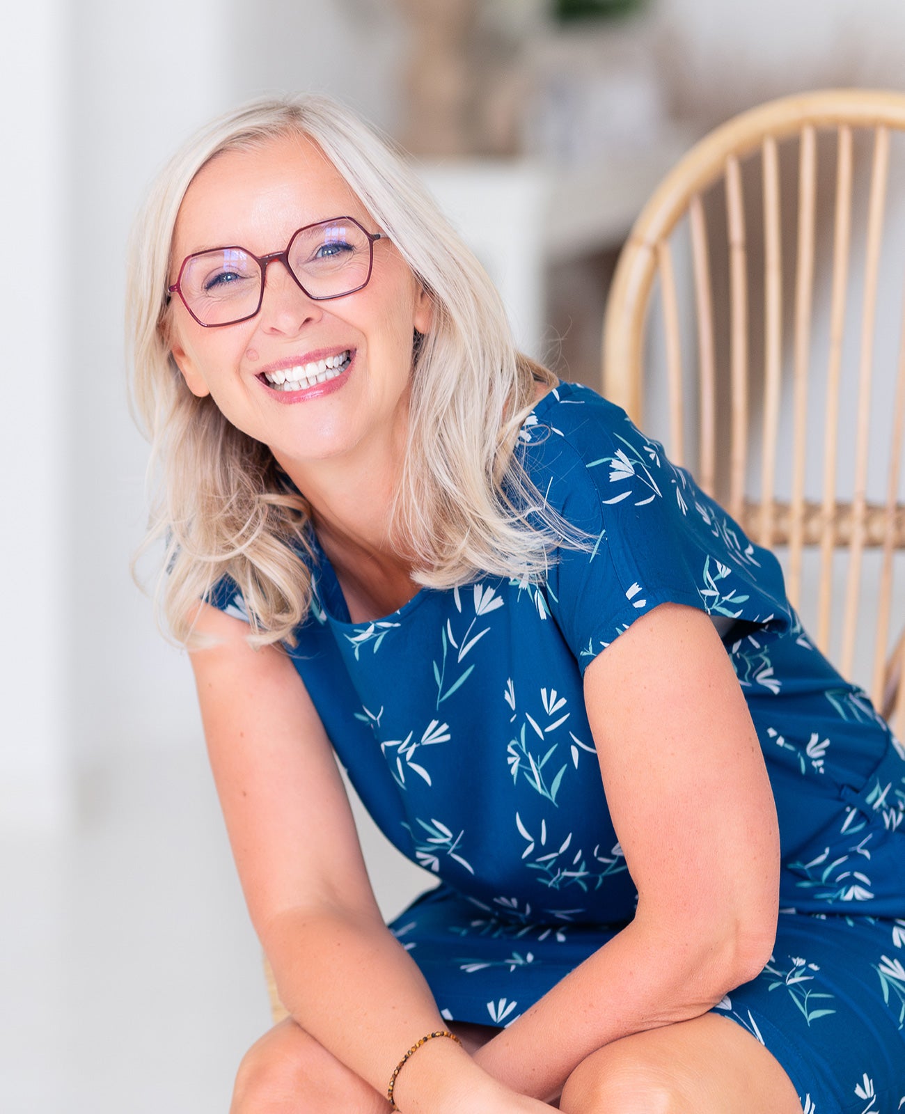 Lächelnde Frau mit blonden Haaren und Brille, trägt blaues Kleid mit floralem Muster, sitzt auf einem Stuhl