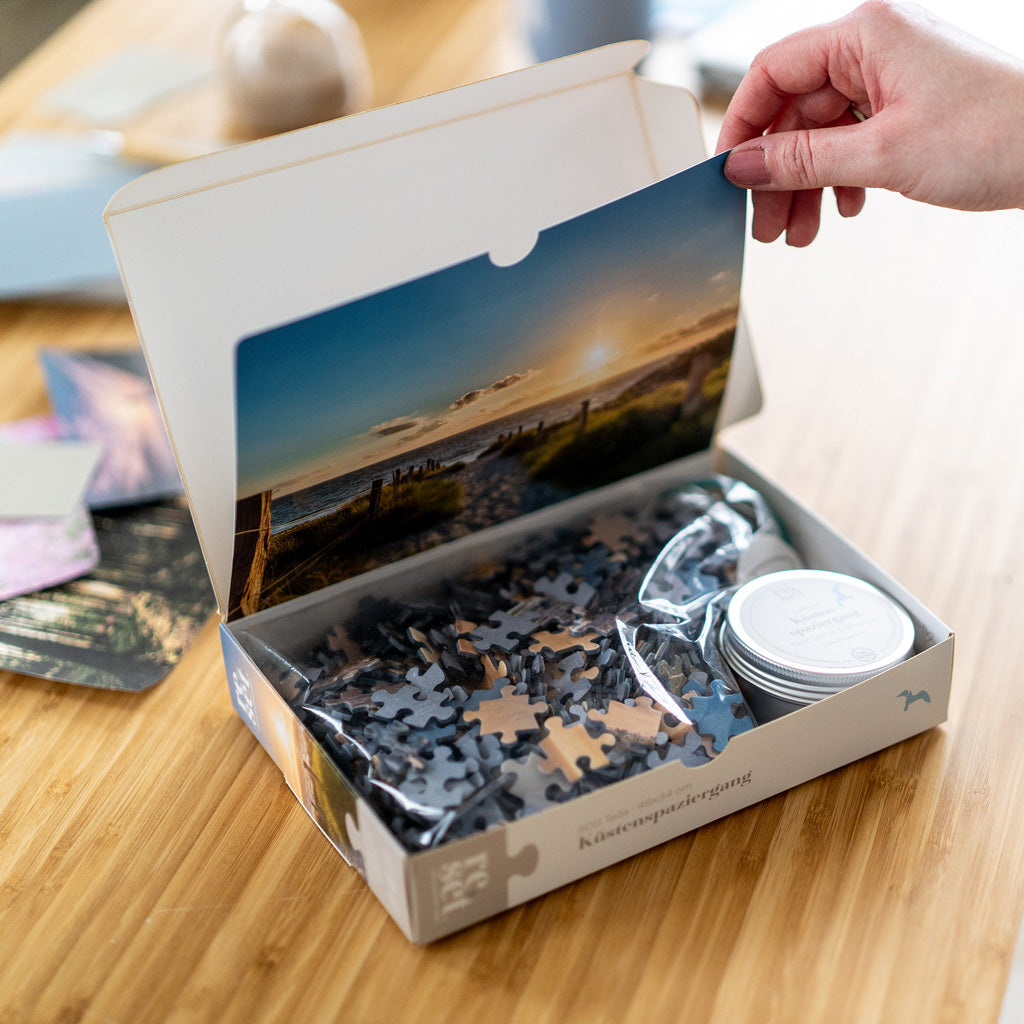 Hand öffnet Puzzleverpackung mit Landschaftsfoto im Deckel, Puzzleteile in Plastiktüte und kleine Dose im Karton