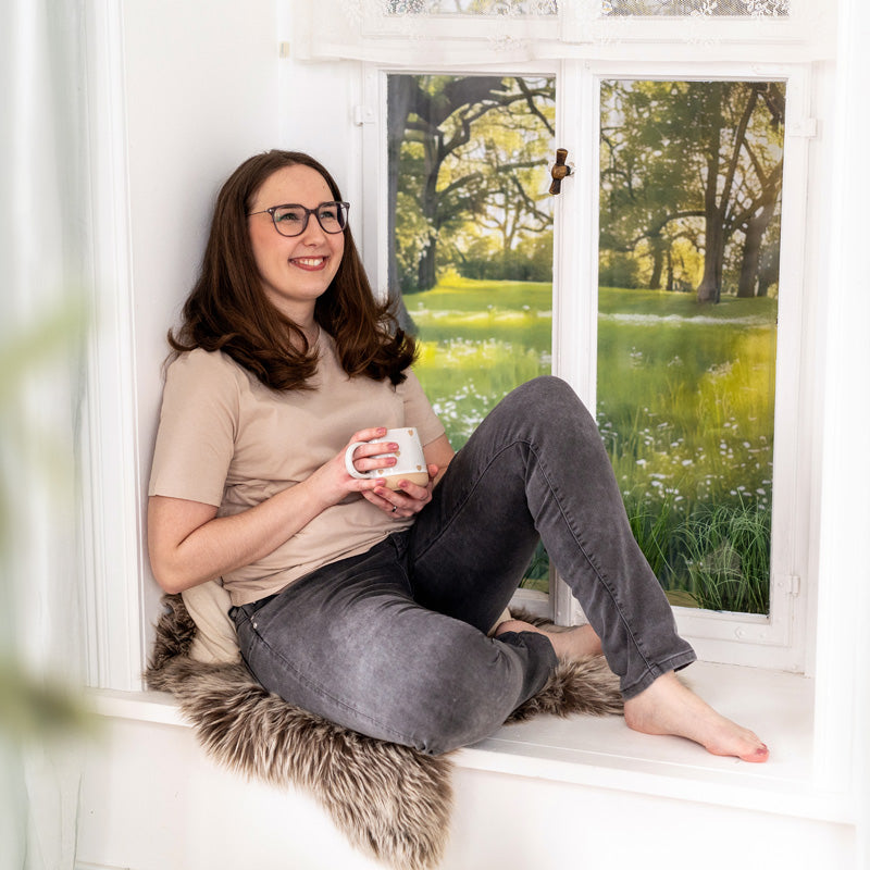 Gründerin des Unternehmens sitzt entspannt auf Fensterbank mit grauer Hose und beigem T-Shirt, hält Tasse, Blick aus Fenster auf grüne Wiese mit Bäumen