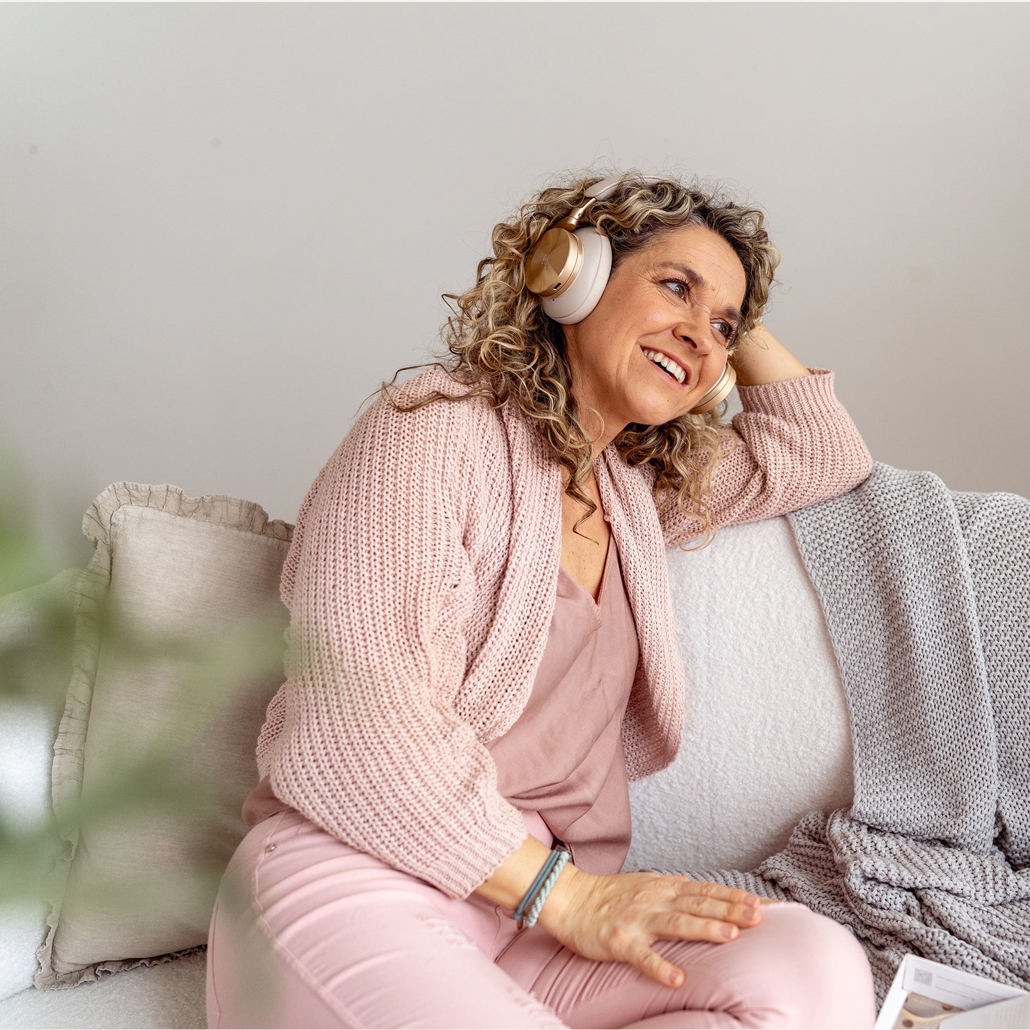 Frau mit lockigem Haar und beigen Kopfhörern lächelt entspannt auf einem Sofa mit rosa Pullover und Hose, grauer Decke und Kissen