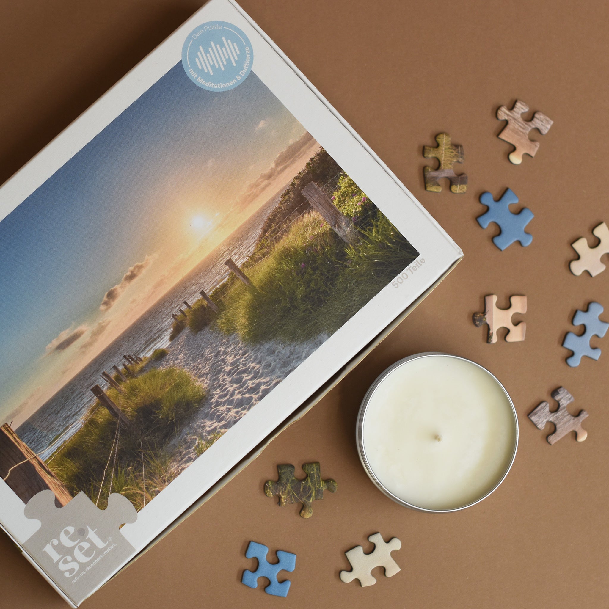 Puzzle mit Sonnenuntergang am Strand, umgeben von einzelnen Puzzleteilen und einer weißen Kerze in Dose auf braunem Untergrund