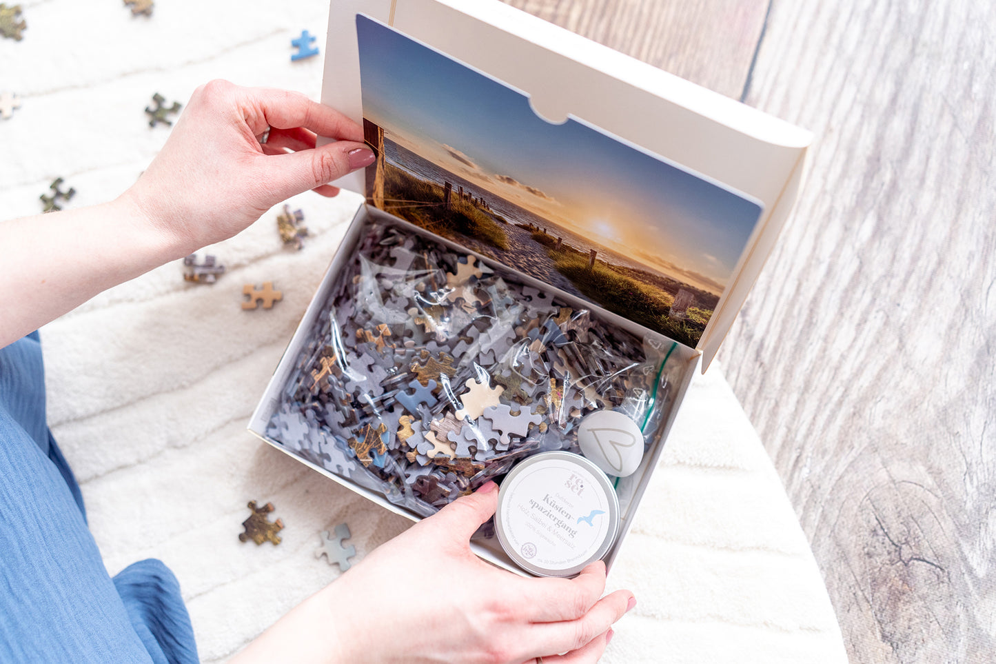 Persön öffnet Puzzlebox mit Meeresmotiv, Duftkerze in einer Dose mit der Aufschrift "Küstenspaziergang" und verstreuten Teilen auf hellem Untergrund.