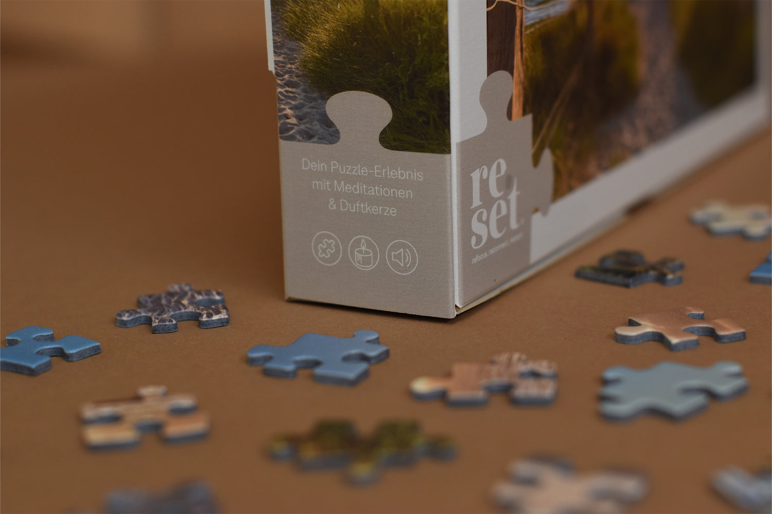 Puzzlebox mit Text 'Dein Puzzle-Erlebnis mit Meditationen & Duftkerze' und verstreute Puzzleteile auf braunem Untergrund