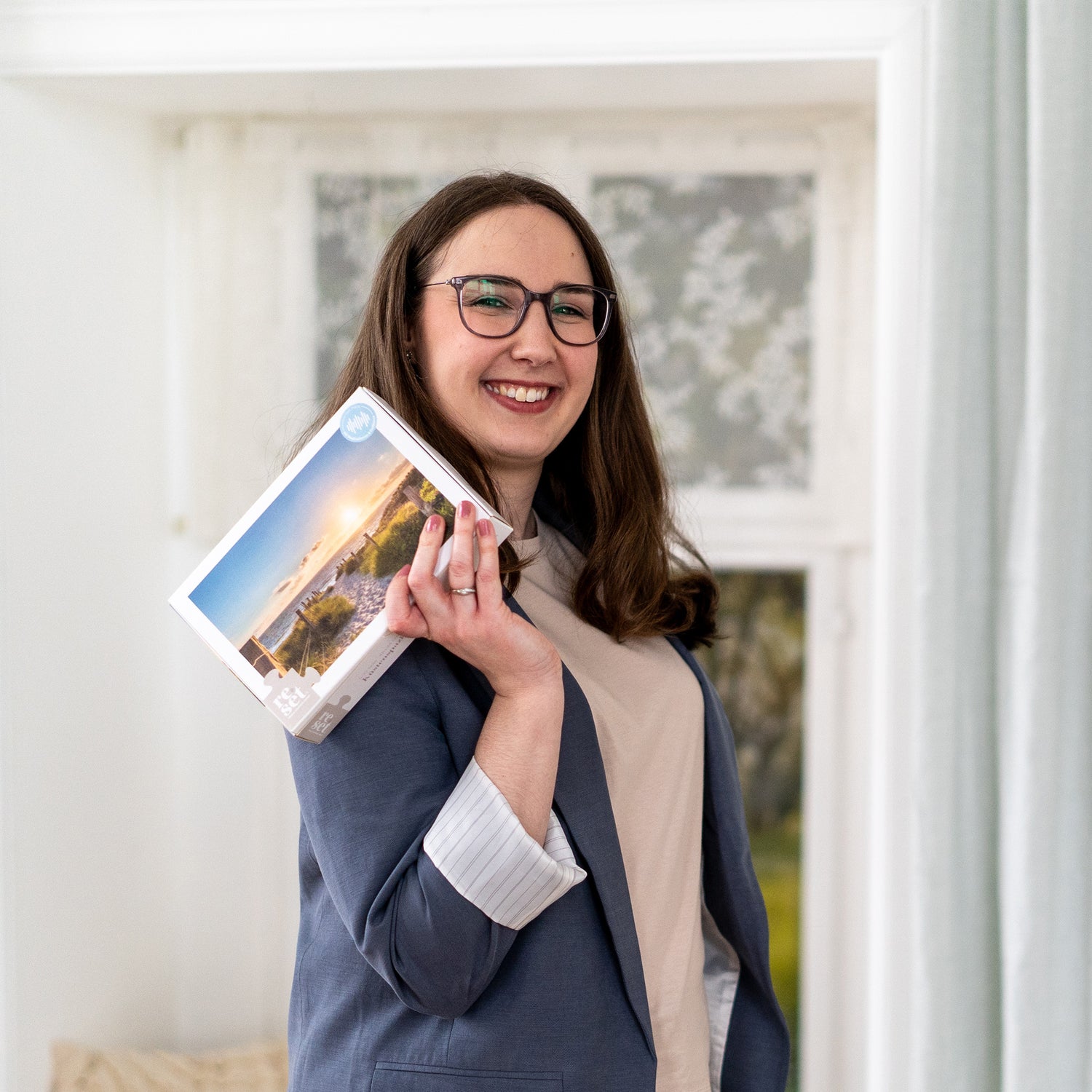 Lächelnde Gründerin mit Brille und blauem Blazer hält eine Puzzle-Schachtel mit Landschaftsbild in der Hand