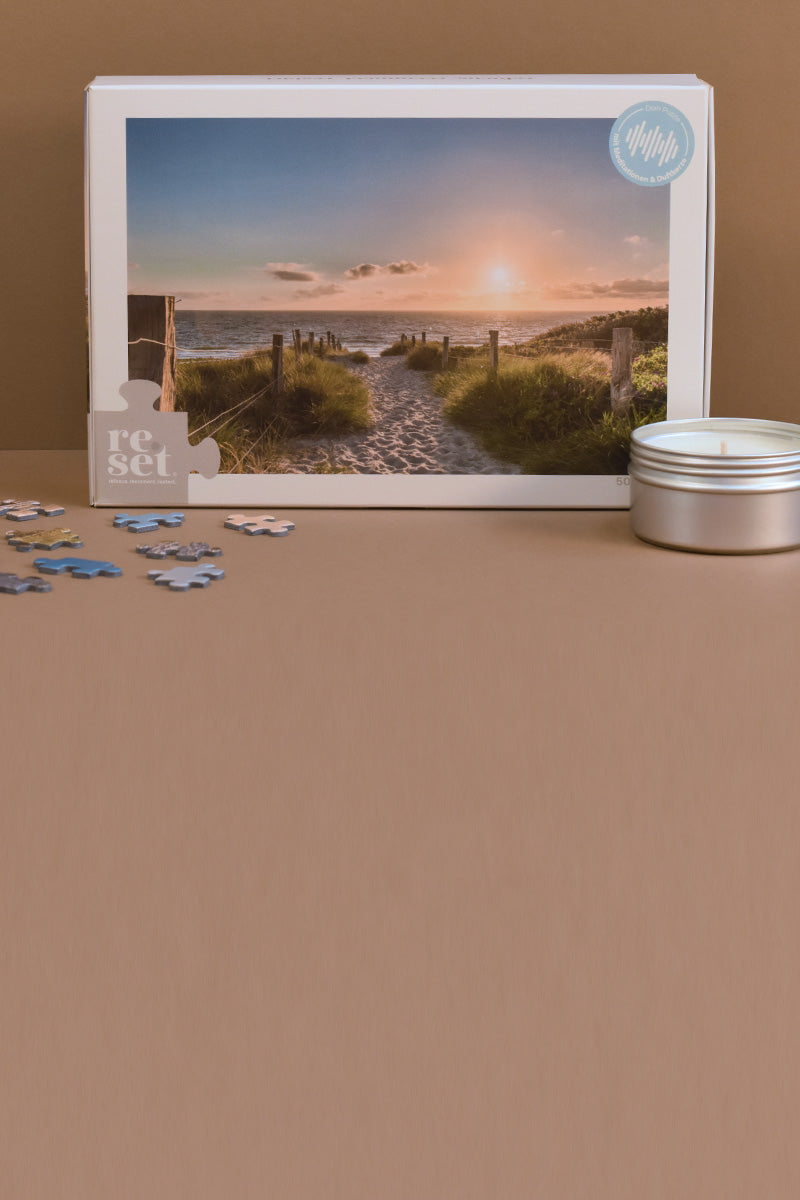 Puzzle mit Motiv eines Strandwegs zum Meer bei Sonnenuntergang, daneben eine kleine weiße Kerze in einer Metallbox und einige lose Puzzlenteile