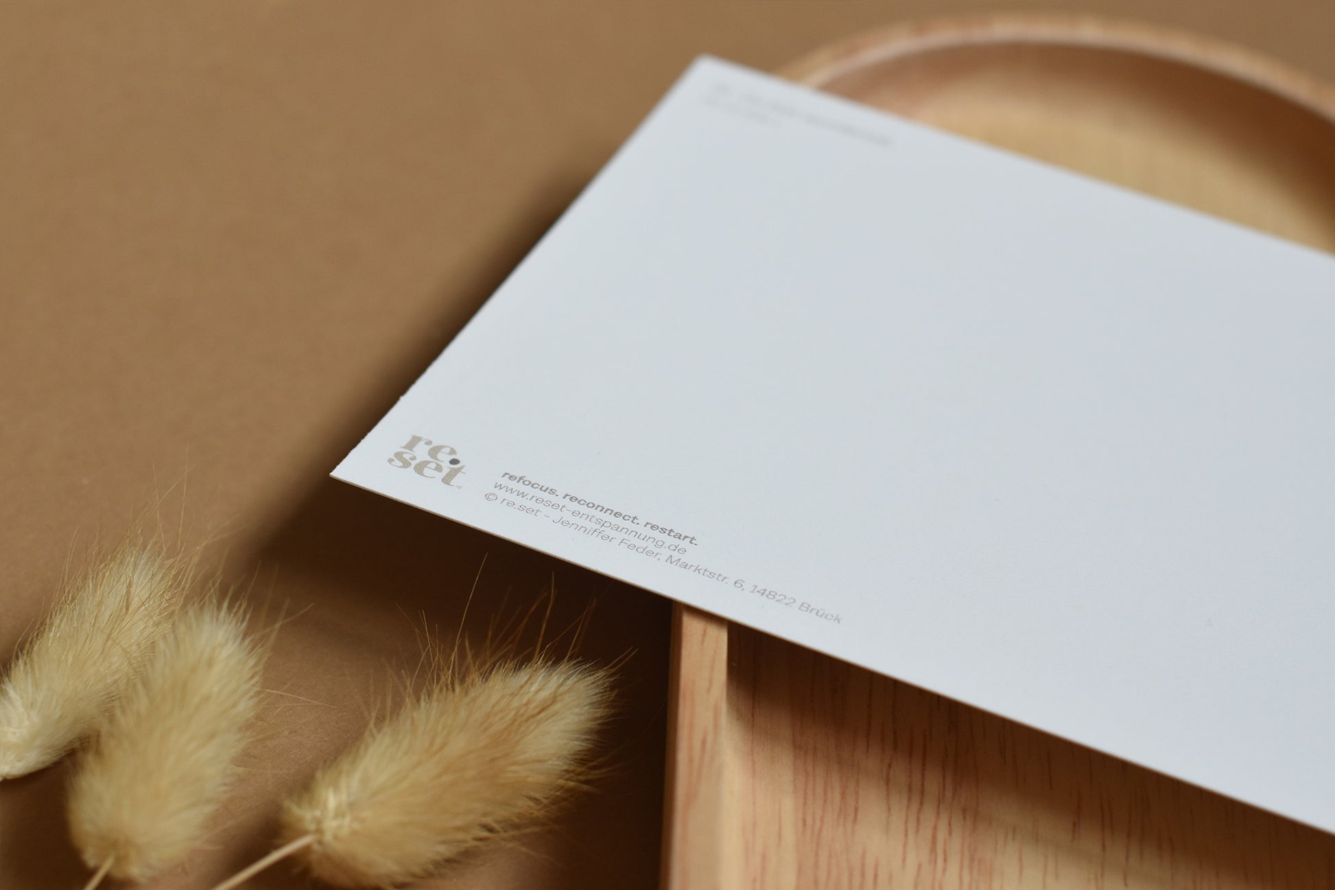 Rückseite einer weißen Postkarte mit kleinem Logo 'reset' auf braunem Untergrund mit getrockneten Gräsern und Holztablett