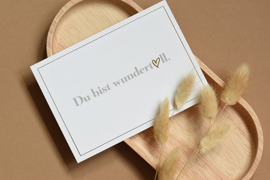 Helle Postkarte mit hellbraunem Text 'Du bist wundertoll' und goldenem Herz, auf einem Holztablett mit getrockneten Blumen