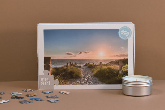Puzzle mit Motiv eines Strandwegs zum Meer bei Sonnenuntergang, daneben eine kleine weiße Kerze in einer Metallbox und einige lose Puzzlenteile