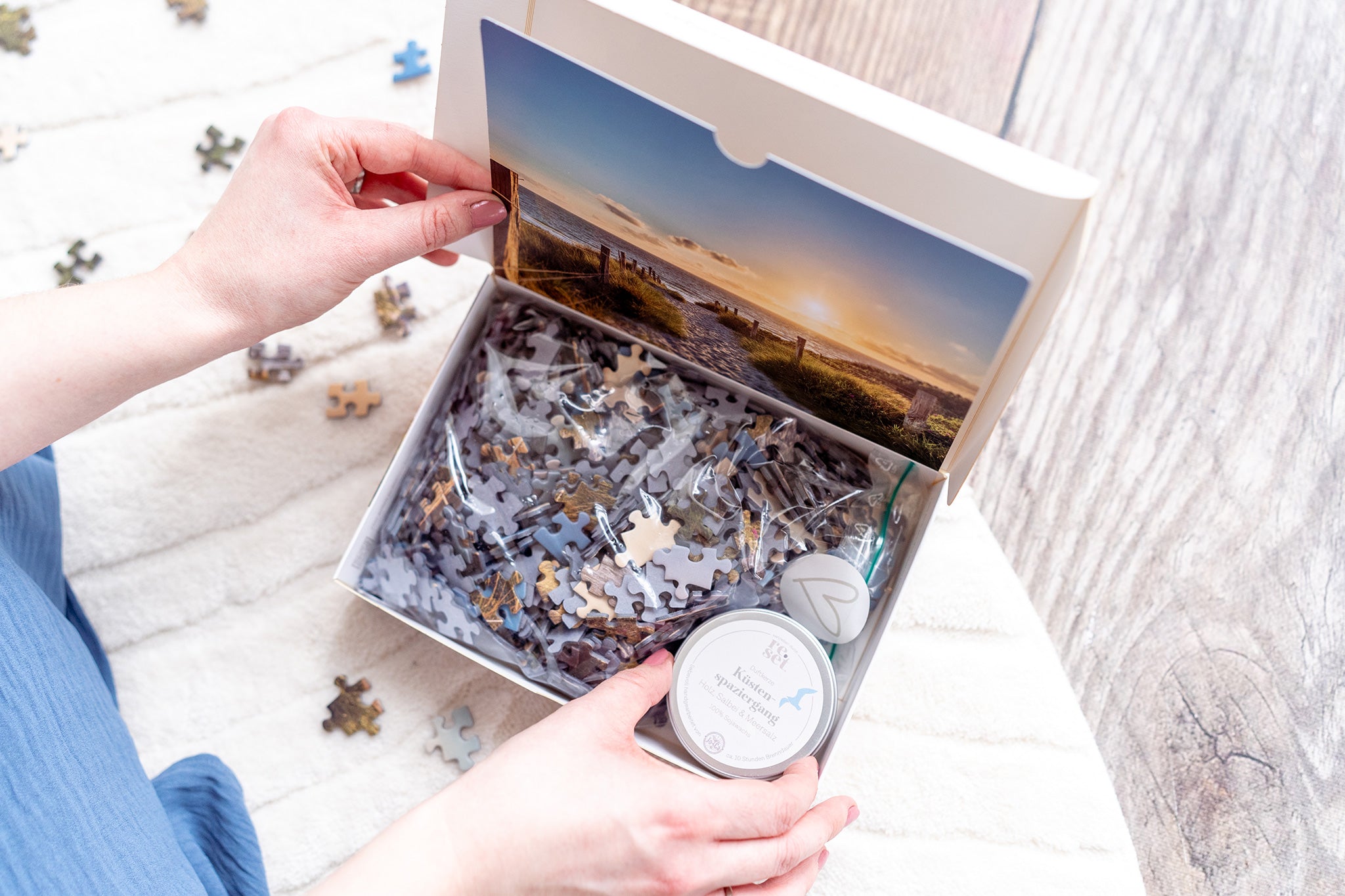 Persön öffnet Puzzlebox mit Meeresmotiv, Duftkerze in einer Dose mit der Aufschrift "Küstenspaziergang" und verstreuten Teilen auf hellem Untergrund.