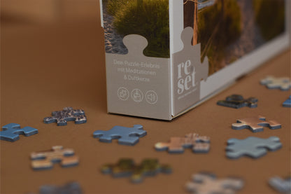 Puzzlebox mit Text 'Dein Puzzle-Erlebnis mit Meditationen & Duftkerze' und verstreute Puzzleteile auf braunem Untergrund