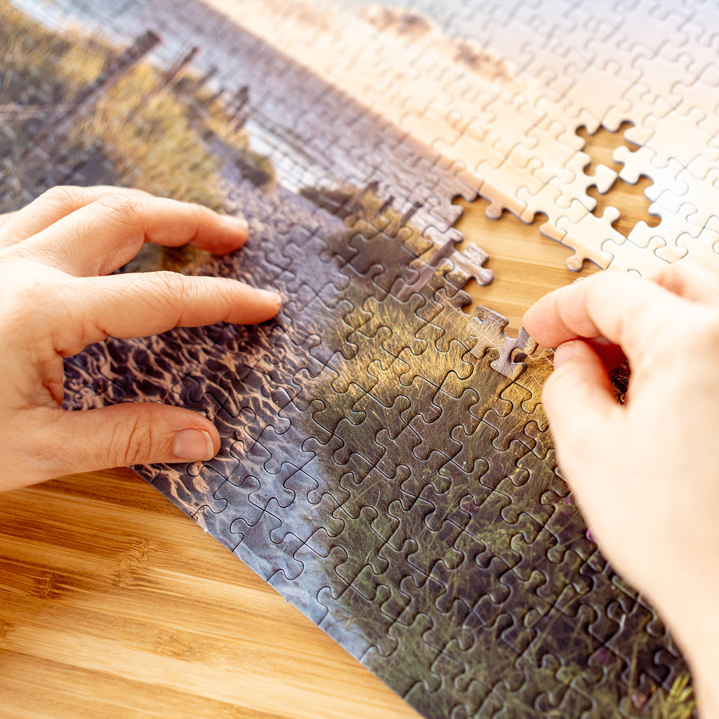 Hände setzen ein fehlendes Puzzlestück in ein Landschaftspuzzle mit Küstenmotiv ein